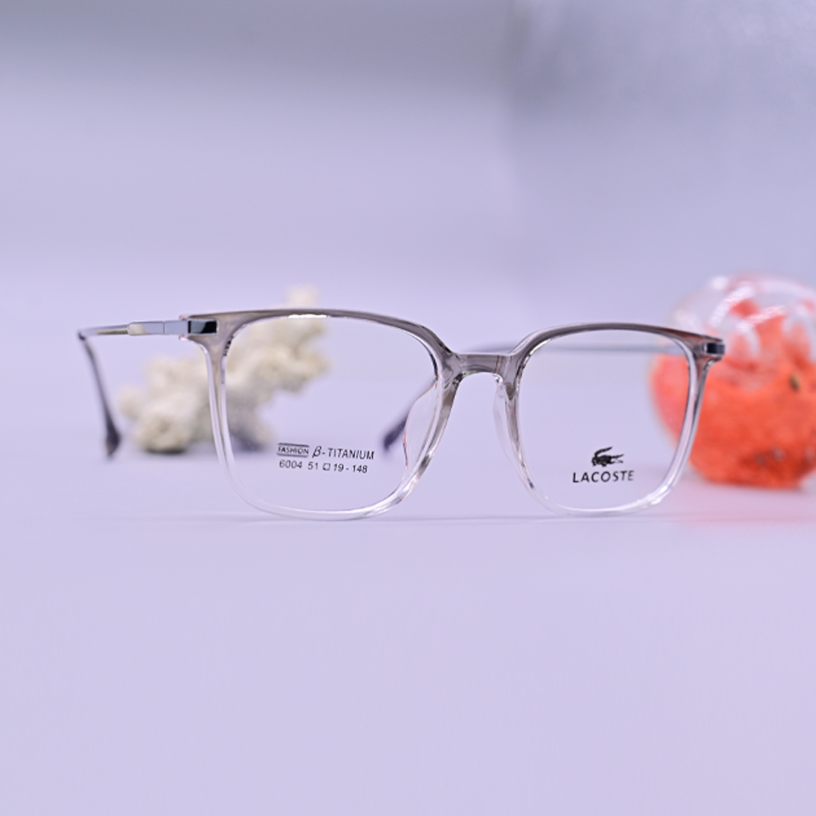 نظارة لاجوست 6004 طبية دائرية بإطار كامل بلون رمادي شفاف وأذرع تحمل شعار العلامة التجارية، تتميز بتصميم كلاسيكي خفيف الوزن وعملي.